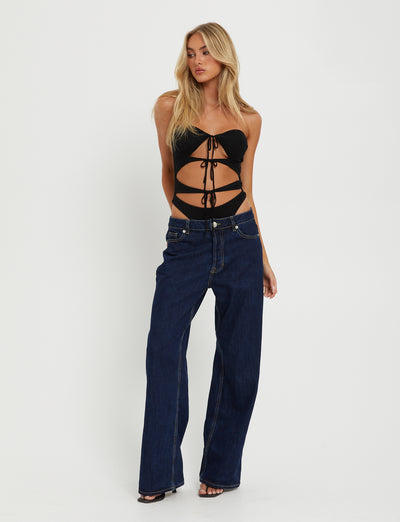 A blonde woman stands against a plain white background, wearing the KINERA BODYSUIT - BLACK from TIGERMIST and blue wide-leg jeans. She has her left hand resting on her thigh and her right hand lightly touching her upper arm.