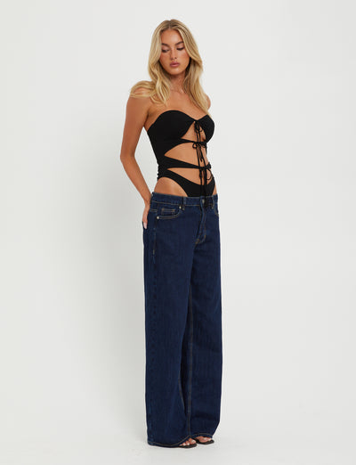 A woman with long blonde hair is modeling the KINERA BODYSUIT in black from TIGERMIST and high-waisted, wide-leg dark blue jeans. She stands against a plain white background, looking slightly down to her left with her hands in the front pockets of her jeans.