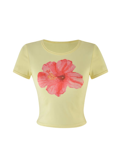 The JOVANA TEE by TIGERMIST is a light yellow, short-sleeved T-shirt with a fitted cut and round neckline. It prominently features a large, vibrant red-pink hibiscus flower design in the center.