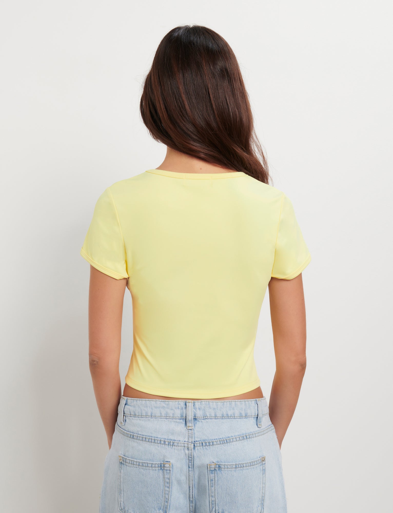 Rear view of a person with long brown hair, wearing the TIGERMIST JOVANA TEE - YELLOW : MACRO FLORAL : HIBISCUS and light blue high-waisted jeans. The individual is standing against a plain white background.
