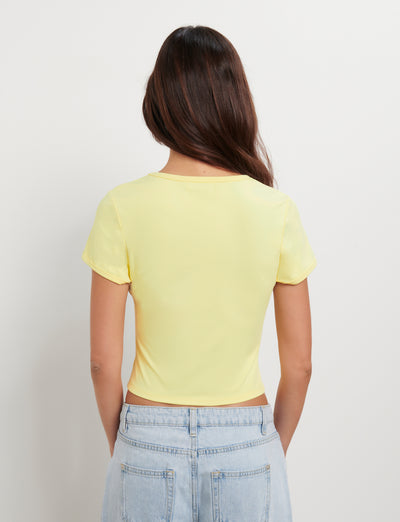 Rear view of a person with long brown hair, wearing the TIGERMIST JOVANA TEE - YELLOW : MACRO FLORAL : HIBISCUS and light blue high-waisted jeans. The individual is standing against a plain white background.
