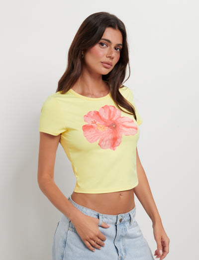 A woman with long, wavy brown hair is wearing the JOVANA TEE in yellow, featuring a large pink hibiscus design in the center. She pairs it with light blue jeans and stands against a plain white background, looking directly at the camera and showcasing the effortless style of TIGERMIST.