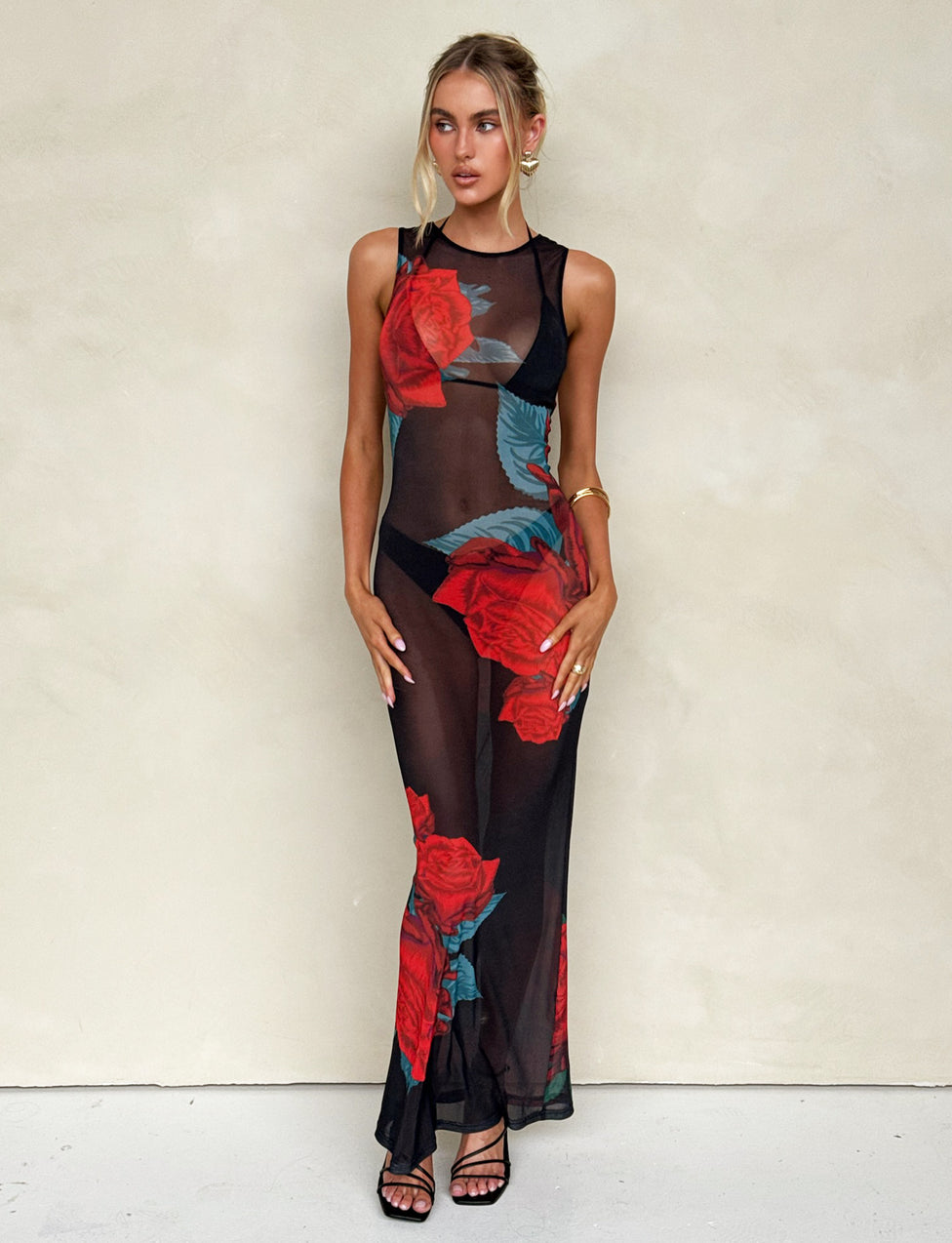 A woman with blonde hair styled in an updo is wearing the JOZIE THREE PIECE DRESS - BLACK : MACRO ROSE - RED ROSE from TIGERMIST, featuring a sheer black fabric adorned with large, vibrant red roses and green leaves. She stands against a plain, light-colored background with her hands resting by her sides, completing the look with black high-heeled sandals.
