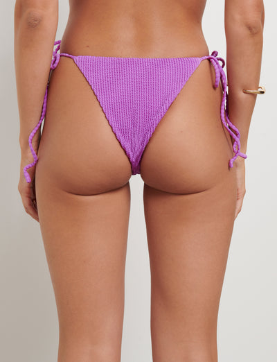A person is shown from the back, wearing the YVE BIKINI BOTTOM in Violet by TIGERMIST. The high-cut, textured bikini bottom features side ties. The skin tone is tan, and an arm bracelet is visible on the right arm against a plain and neutral background.