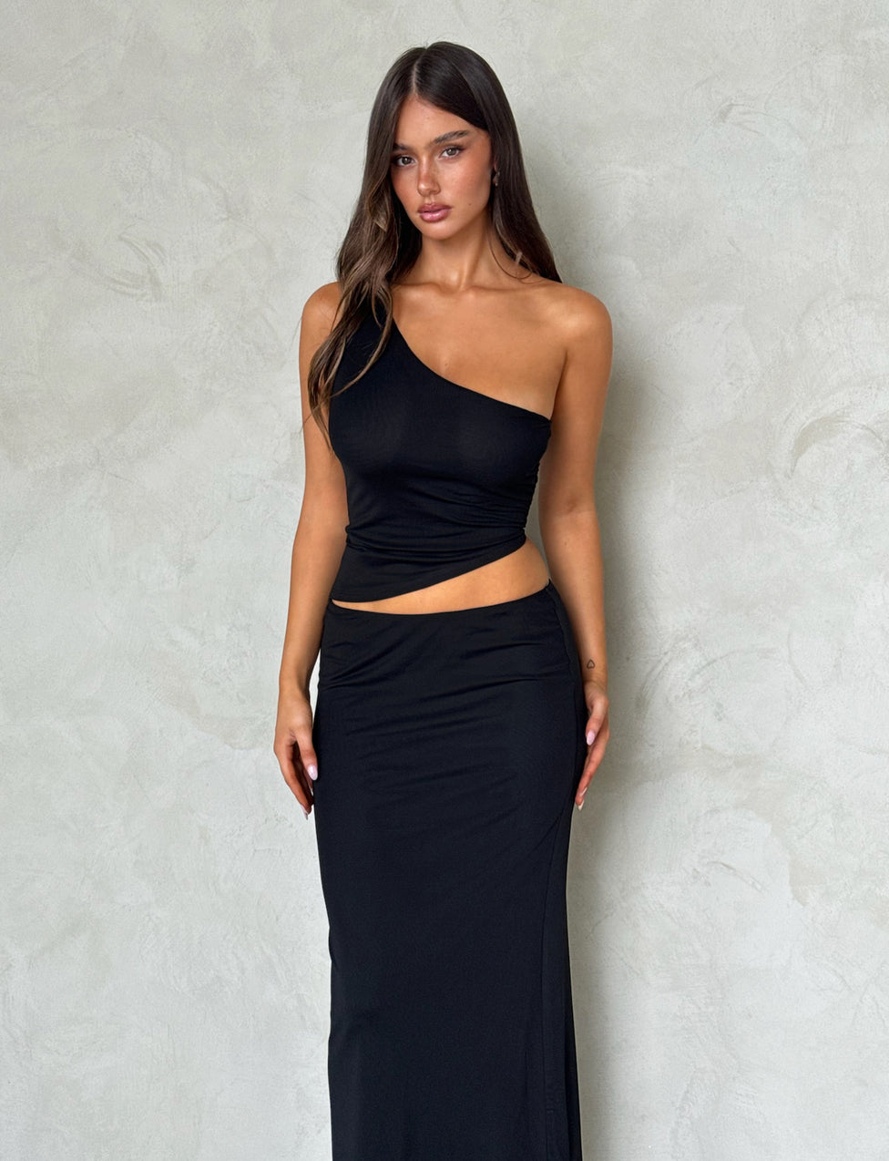 A woman with long, brown hair is wearing a stylish black DEANNA TOP from TIGERMIST and a matching Tiger Mist long skirt. She stands against a textured light gray background, looking directly at the camera with a neutral expression.