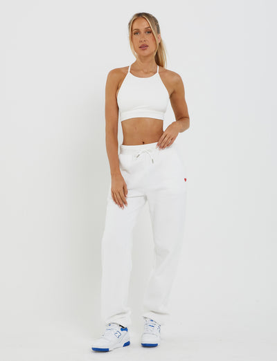 A woman stands against a white background wearing a white sports bra, the TIGERMIST LUMEN PANT in white, featuring a mid-rise waist with drawstring and an oversized trackpant silhouette, and white sneakers with blue accents. She has her left hand in her pocket and her right arm relaxed by her side.