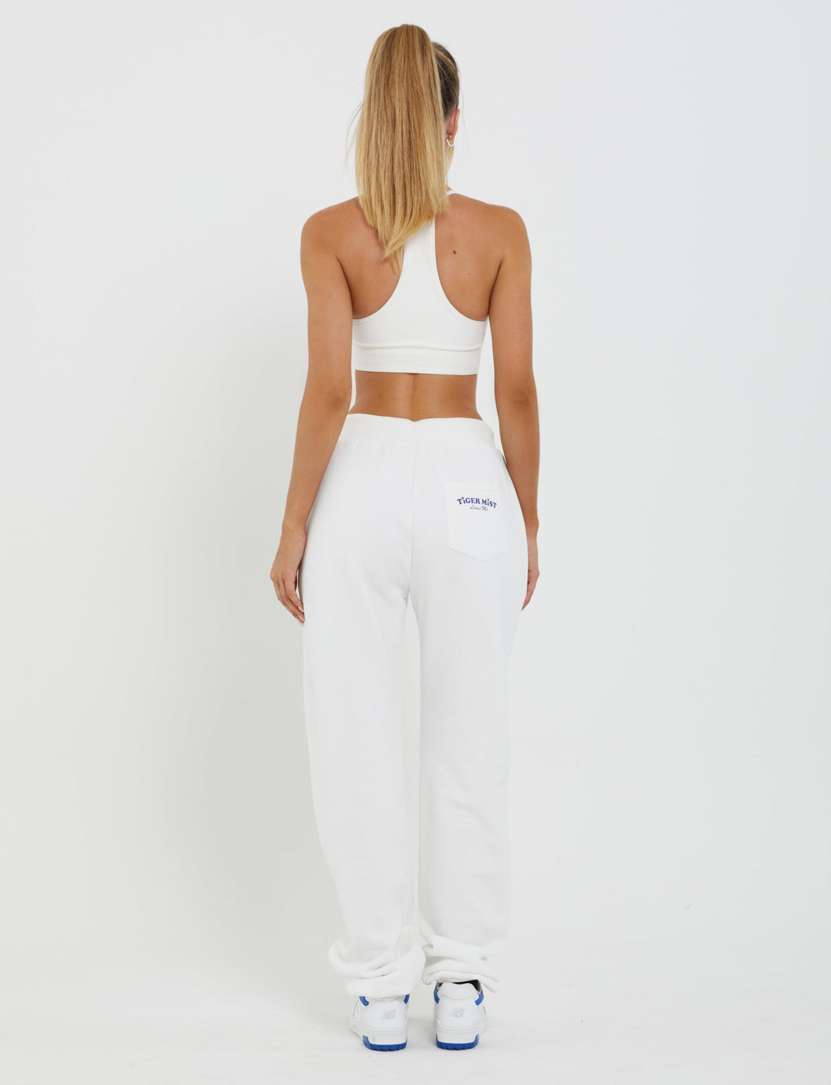 A woman stands with her back to the camera, dressed in the TIGERMIST LUMEN PANT - WHITE and a matching white sports bra. She has long blonde hair styled in a high ponytail and is positioned against a plain white backdrop. Blue text adorns the back pocket of her mid-rise loungewear pants. Her outfit is completed with white sneakers featuring blue accents.