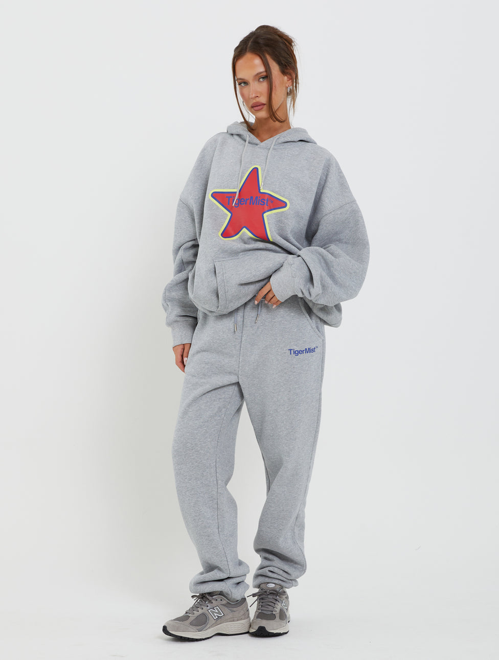 A person stands against a white background wearing a baggy gray hoodie adorned with a colorful star design and the LUMEN PANT - GREY from TIGERMIST. They have one hand in their pocket and the other on their hip, completing the look with gray sneakers.