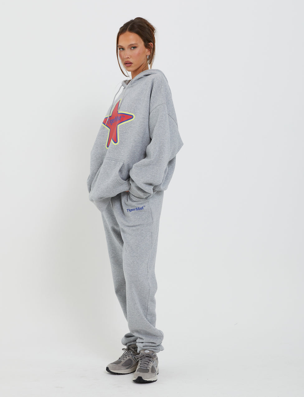 A woman stands wearing an oversized TigerMist Lumen Hoodie in grey, paired with matching sweatpants, her hands in the pockets. The hoodie features a large red star design on the front. She is also wearing grey athletic shoes and has her hair tied back while looking at the camera with a neutral expression.