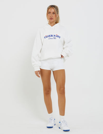 A woman stands against a plain white background, dressed in an oversized LUMEN HOODIE - WHITE from TIGERMIST with "TIGER MIST Loves Me" written on it. She pairs the hoodie with white shorts and white sneakers featuring blue detailing. Her hair is styled in a ponytail, and her hands rest casually by her sides.