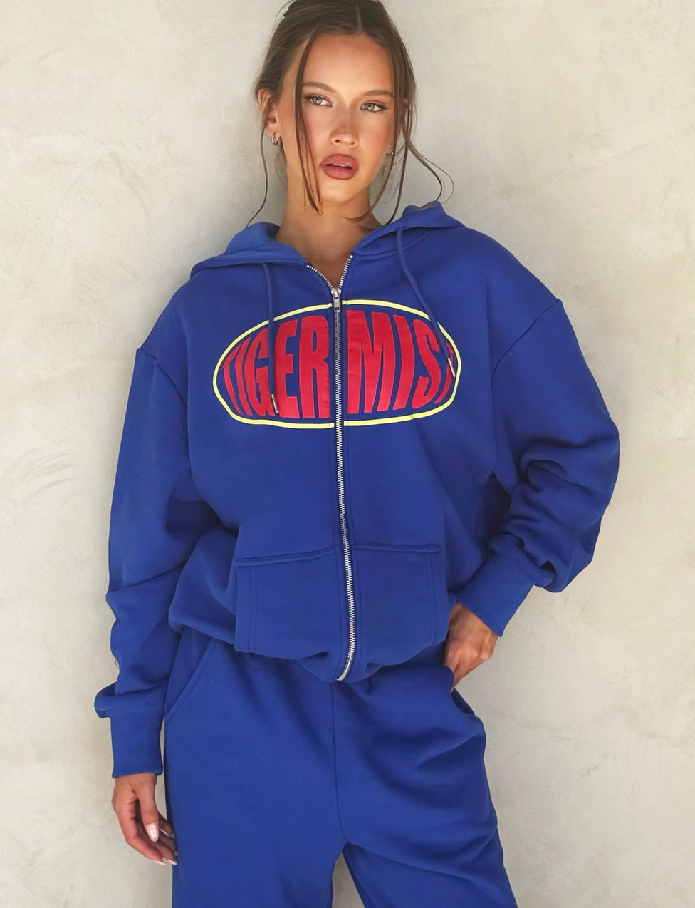 A person wearing the LUMEN ZIP FRONT HOODIE - BLUE from TIGERMIST stands confidently against a light-colored background. Paired with matching blue sweatpants, the zip-up hoodie enhances their relaxed yet stylish look, accentuated by their hand placed coolly on their hip.