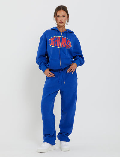 A woman stands against a plain white background wearing the LUMEN ZIP FRONT HOODIE - BLUE from TIGERMIST and matching blue drawstring sweatpants. She is also wearing white sneakers. Her hands are placed in her pants pockets, and she has a confident expression.