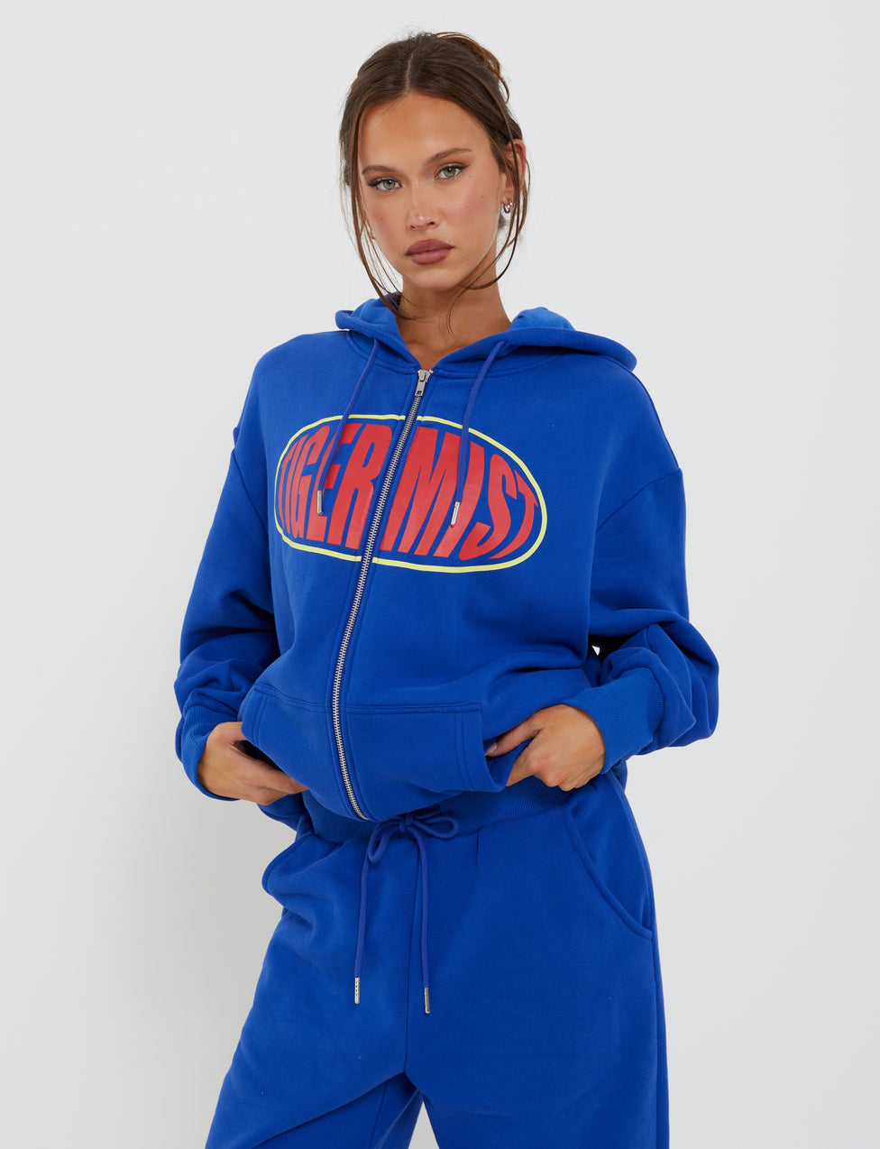 A woman stands against a plain background wearing the LUMEN ZIP FRONT HOODIE - BLUE from TIGERMIST, with "MERMS" printed in bold red letters on the front. She has her hands in the hoodie pockets and is also wearing matching blue sweatpants. Her hair is styled with a middle part and tied back.