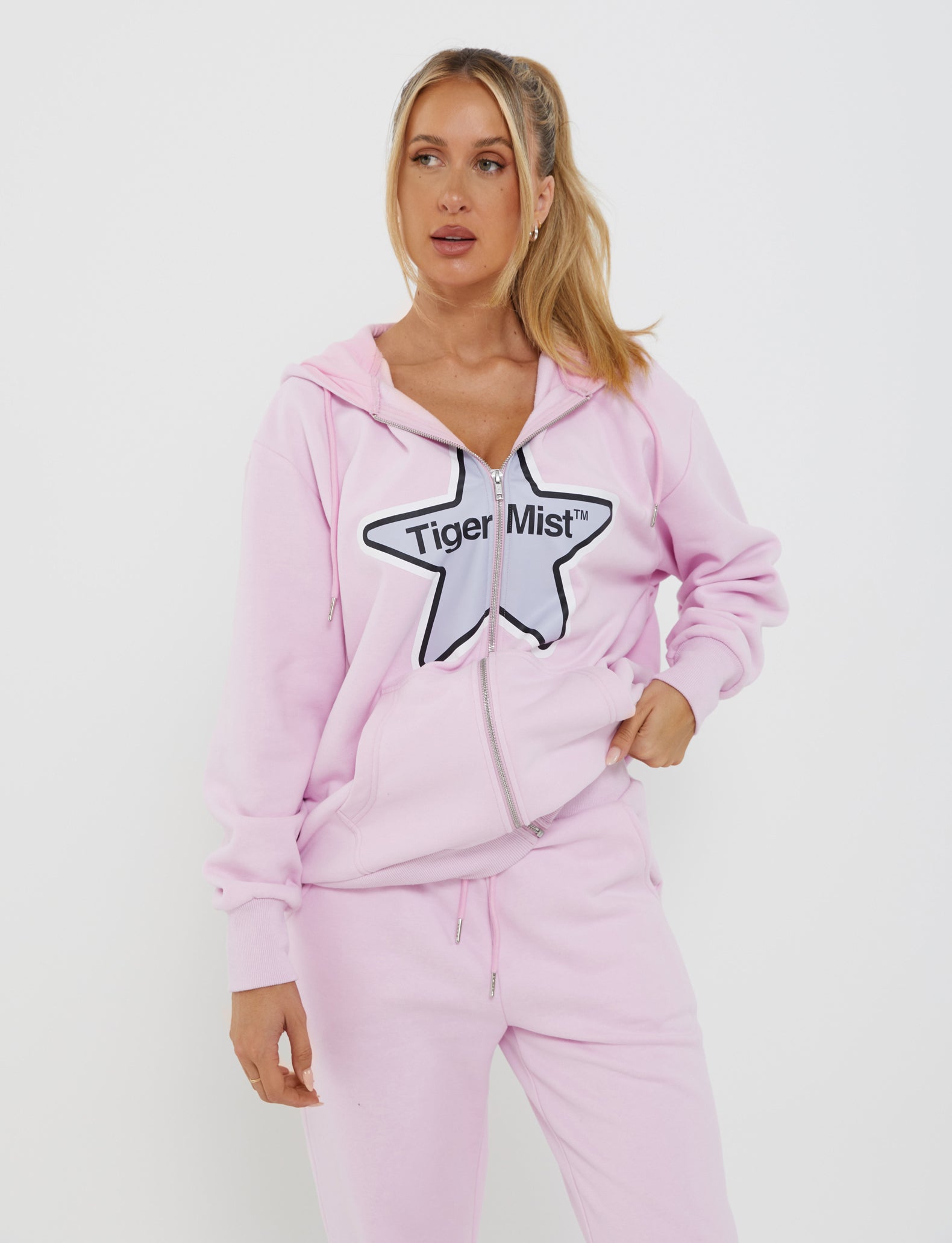 A woman with long blonde hair in a ponytail wears an oversized LUMEN ZIP FRONT HOODIE - PINK from TIGERMIST. She pairs it with matching fleece-lined sweatpants, standing against a plain white background, gazing off to the side.