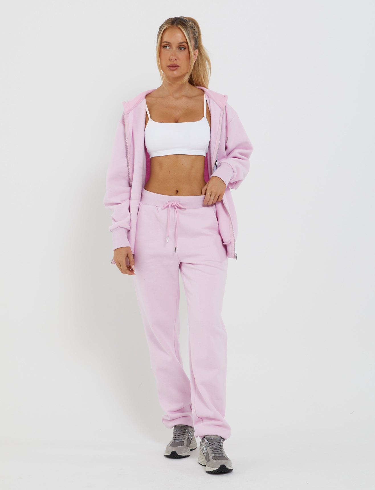 A person wearing the TIGERMIST LUMEN ZIP FRONT HOODIE in pink, paired with matching drawstring sweatpants, a white sports bra, and gray sneakers stands against a plain white background. With one hand casually in their pocket and a relaxed posture, they showcase the adjustable fit of their comfortable outfit.