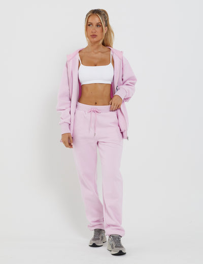 A person wearing the TIGERMIST LUMEN ZIP FRONT HOODIE in pink, paired with matching drawstring sweatpants, a white sports bra, and gray sneakers stands against a plain white background. With one hand casually in their pocket and a relaxed posture, they showcase the adjustable fit of their comfortable outfit.