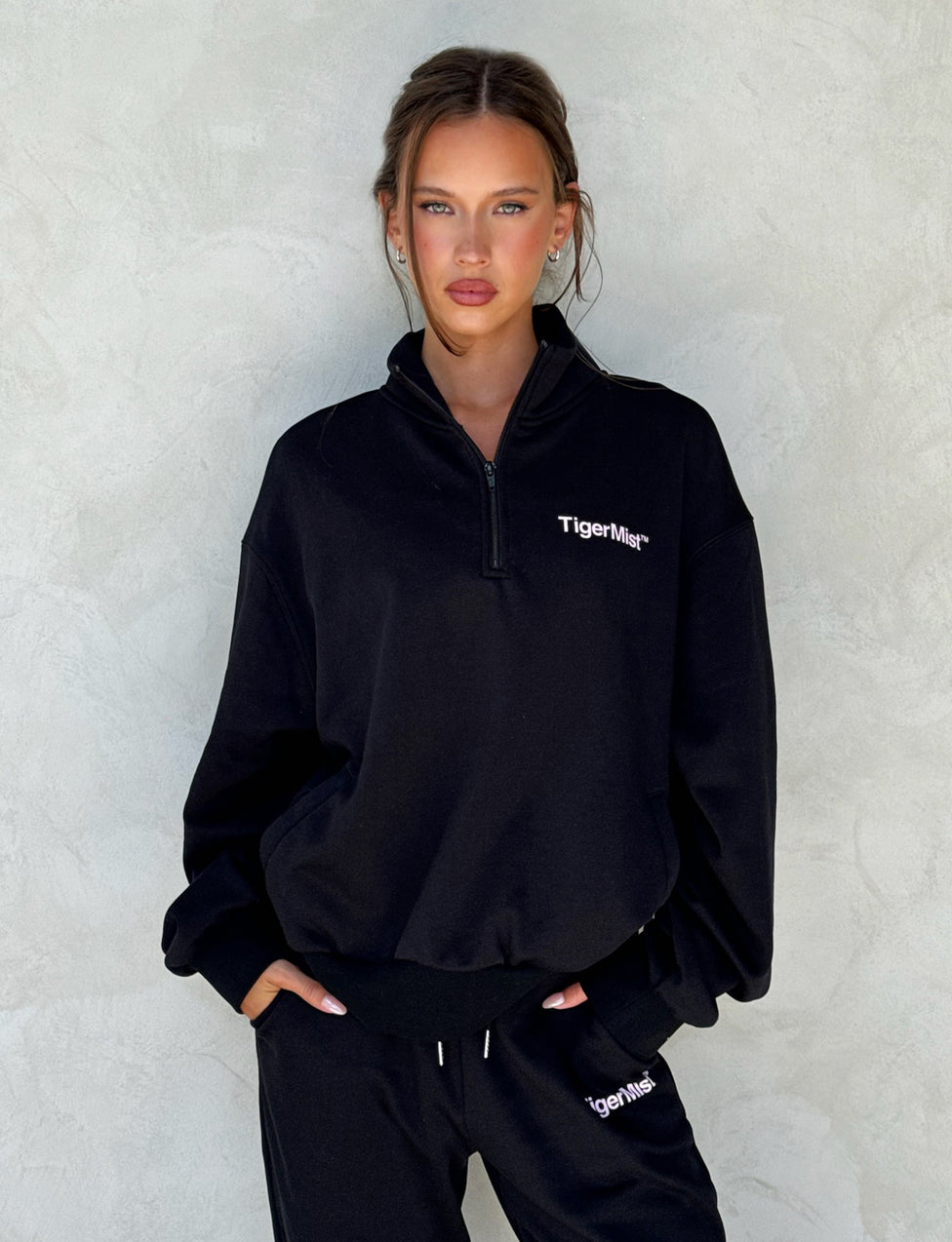 A woman stands against a plain textured background, wearing the loose-fitting DELFINA JUMPER in black from TIGERMIST and matching black pants. She has long hair tied back and her hands are in her pockets, looking directly at the camera with a neutral expression.