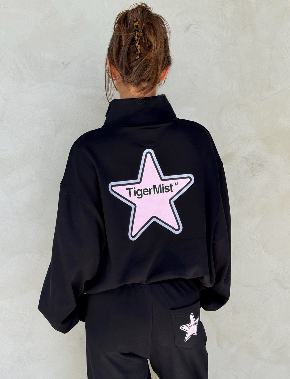 A person with a bun hairstyle is seen from behind wearing an outfit from TIGERMIST. They are sporting the DELFINA JUMPER in black, which features a large pink star emblem with the text "TIGERMIST™" on the back. The left pocket of their matching black pants also has a smaller pink star emblem, showcasing the signature Tiger Mist style.
