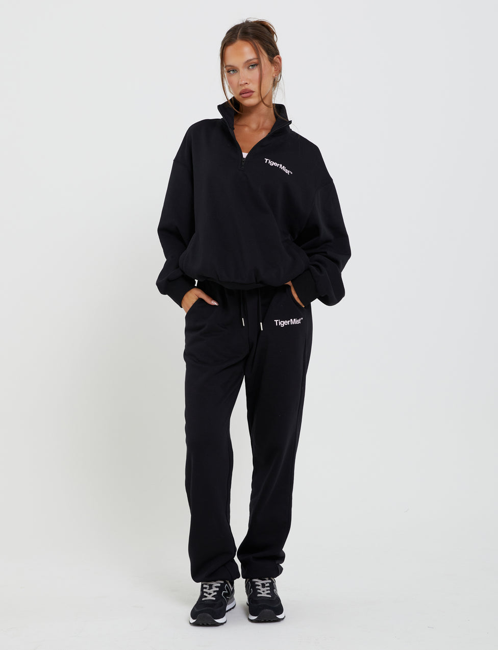 A woman stands against a plain white background wearing the TIGERMIST DELFINA PANT - BLACK tracksuit, which has "TIGERMIST" printed on the top left of both the jacket and pants. She has her hands in her pockets and is also wearing black athletic shoes. Her hair is tied back neatly.