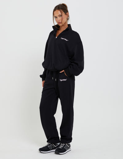 A person stands wearing the DELFINA JUMPER - BLACK and matching black sweatpants, both emblazoned with "TIGERMIST." With hands in pockets and black sneakers completing the look, they pose confidently against a plain white background.