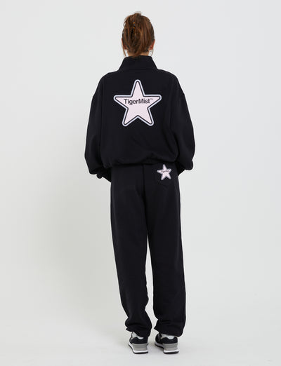 A person with brown hair tied up is standing with their back to the camera, wearing the TIGERMIST DELFINA JUMPER in black. The tracksuit features a large white star on the back with the text "TIGERMIST" inside. They are standing against a plain white background.