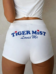 A person is shown from the waist down wearing the DELANEY SHORT in white. The back of the shorts features bold, blue text that reads "TIGERMIST Loves Me," with small red hearts adorning the letters "i" in both "TIGER" and "MIST." Complementing Tiger Mist's playful style, the individual pairs these shorts with a white top.