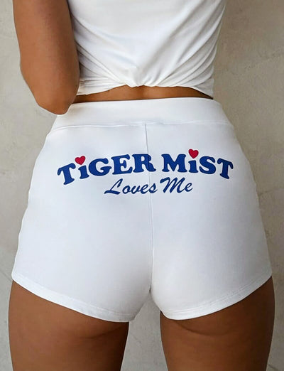 A person is shown from the waist down wearing the DELANEY SHORT in white. The back of the shorts features bold, blue text that reads "TIGERMIST Loves Me," with small red hearts adorning the letters "i" in both "TIGER" and "MIST." Complementing Tiger Mist's playful style, the individual pairs these shorts with a white top.