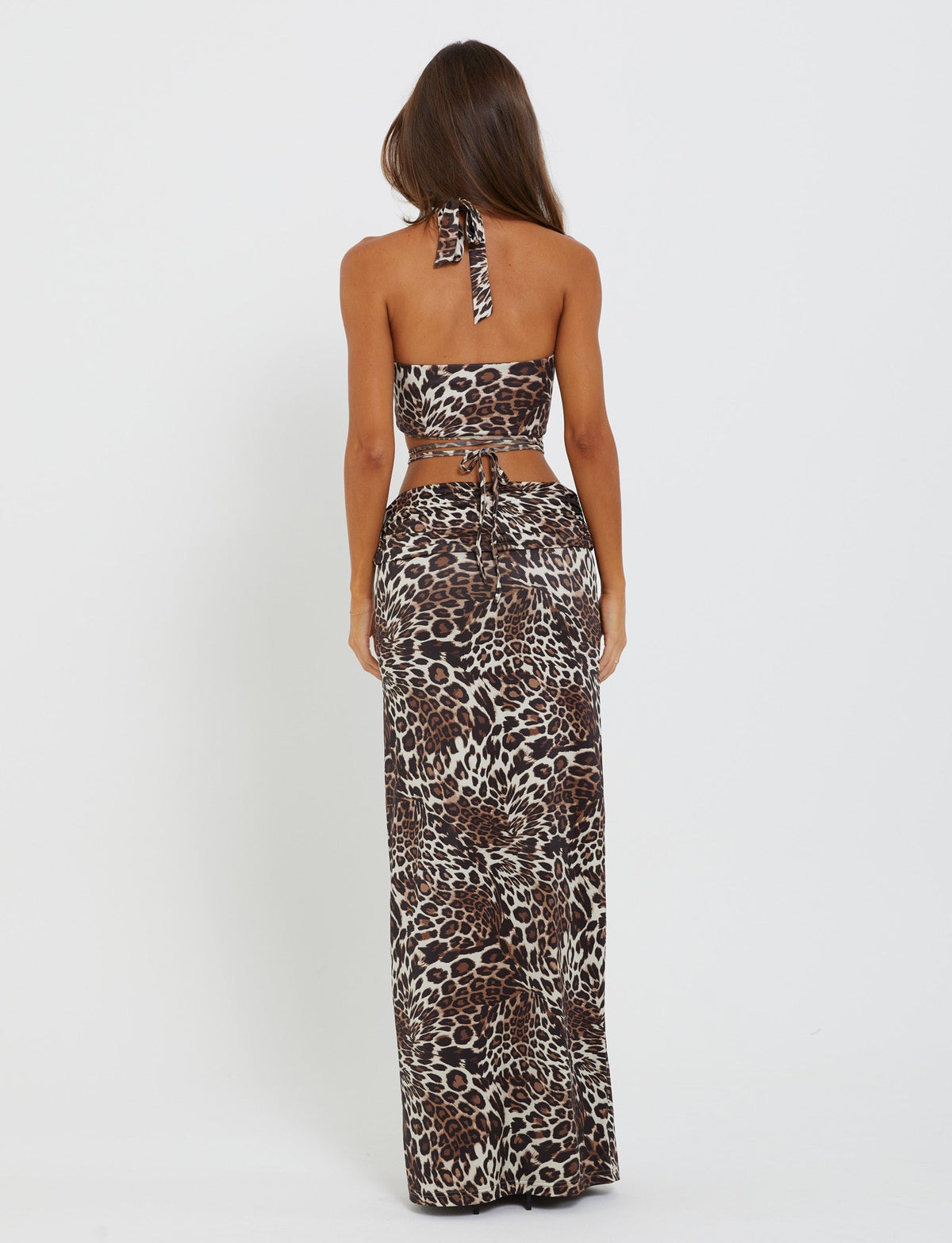A woman with long brown hair is standing with her back to the camera, showcasing her TigerMist halter neck crop top and CYNTHIA MAXI SKIRT - MULTI : LEOPARD. The background is white and plain.
