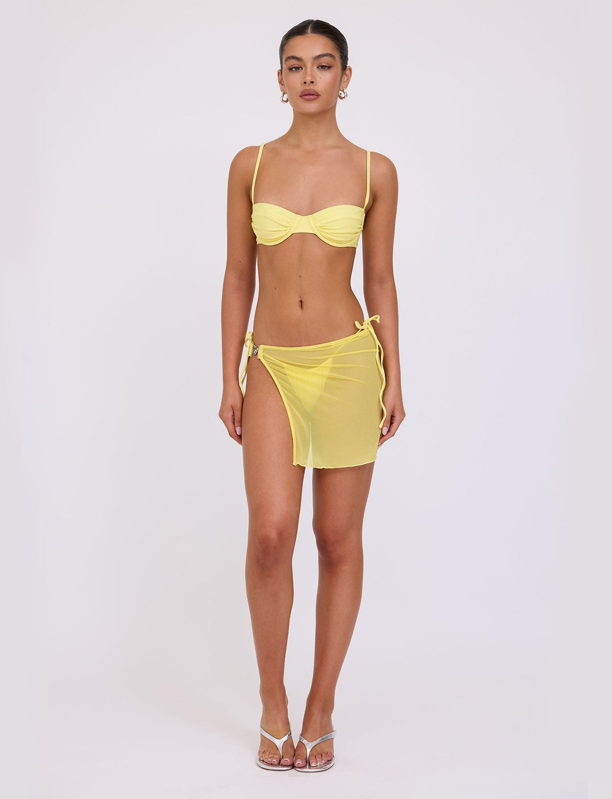 A woman stands against a plain white background, wearing a yellow TIGERMIST bikini top with thin straps and the CYNTHIA MINI MESH SKIRT in pale yellow, tied at the waist to reveal matching bikini bottoms. She has her hair tied back, accessorizes with earrings, and wears clear high-heeled sandals.
