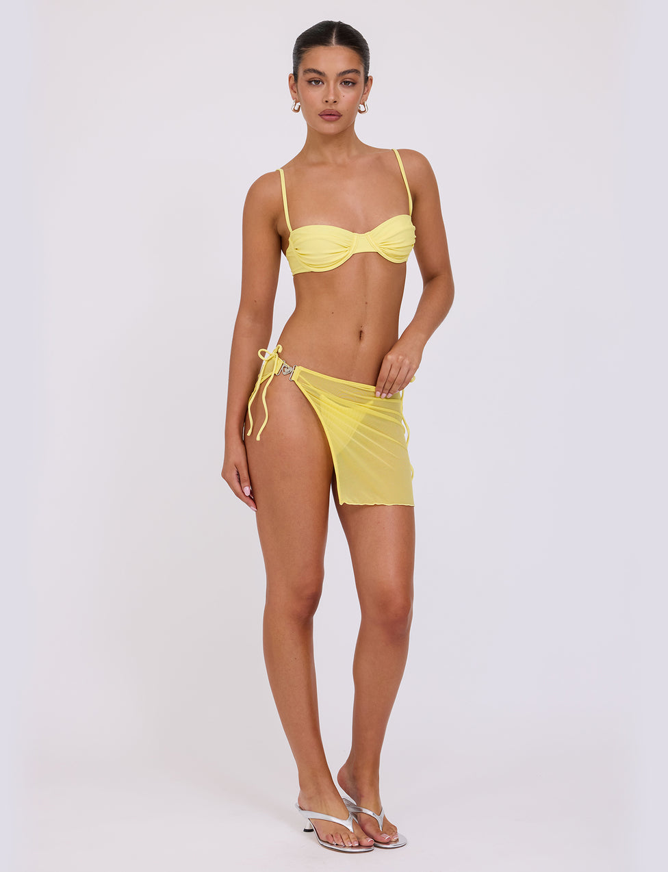 A woman stands against a plain white background wearing a yellow bikini and the CYNTHIA MINI MESH SKIRT in Pale Yellow from TIGERMIST. She has her hair tied back, wears earrings, and is standing in a confident pose with one arm by her side and the other placed on her hip. She is also wearing white sandals.
