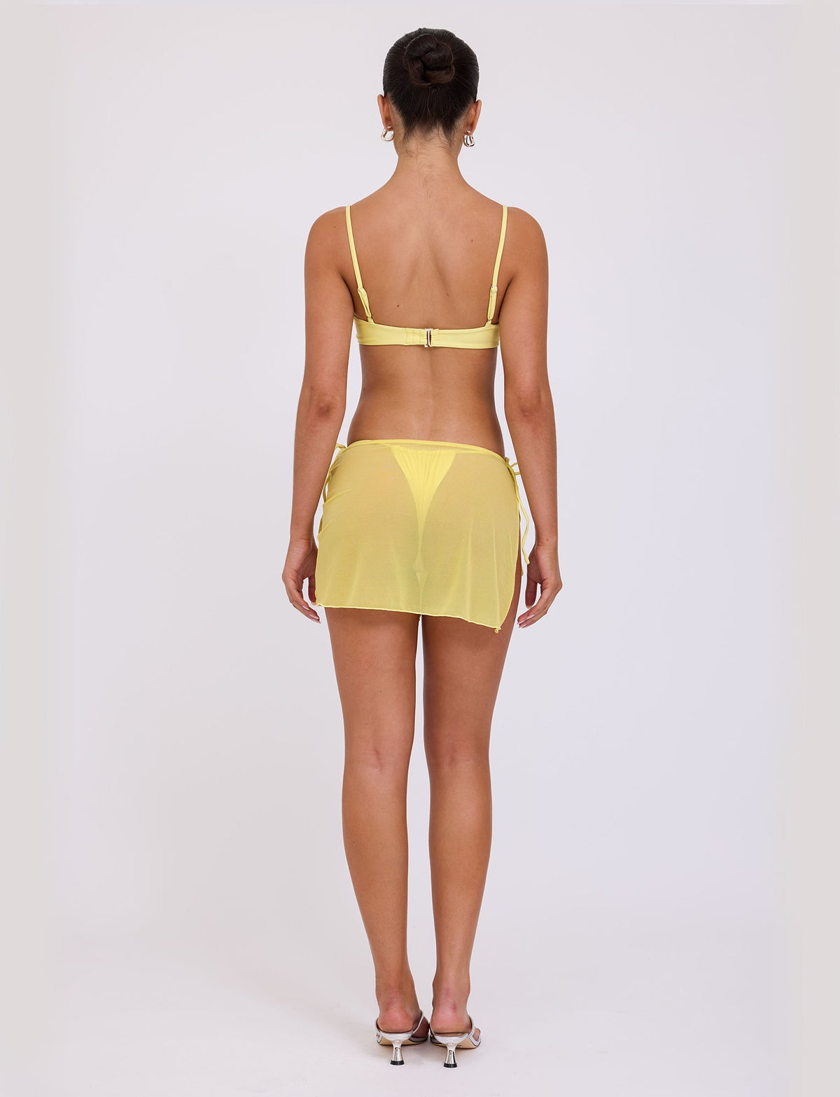 A woman is posing with her back to the camera, styled in a chic TIGERMIST Tiger Mist yellow bikini top and a matching CYNTHIA MINI MESH SKIRT - YELLOW: PALE YELLOW. Her hair is elegantly styled in a bun, and she stands confidently on high-heeled shoes against a plain white background.