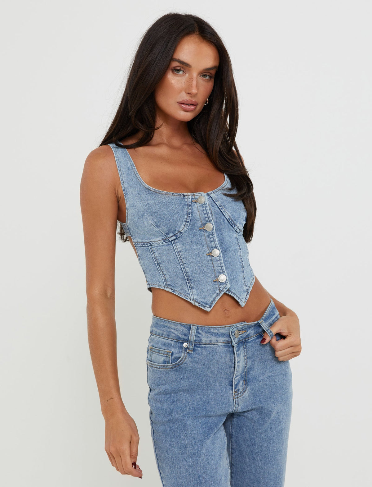 A woman with long, dark hair, dressed in a fitted, sleeveless Tigermist Tatyana Top in light wash light denim adorned with button-down details, pairs it effortlessly with high-waisted jeans. She poses against a plain white background, one hand resting on her hip while the other holds the waistband of her jeans.
