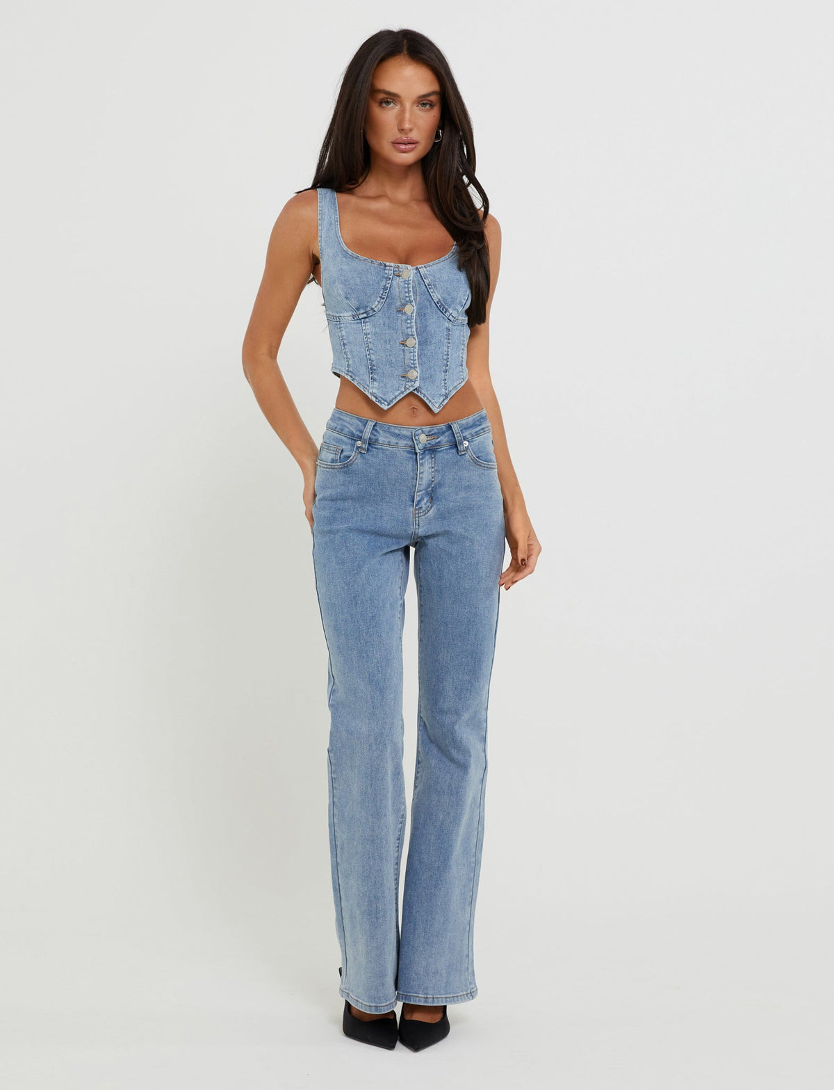A person with long dark hair poses against a plain white background, dressed in a stylish TIGERMIST TATYANA TOP in light wash denim and high-waisted, straight-leg jeans. The ensemble is completed with sophisticated black pointed heels.