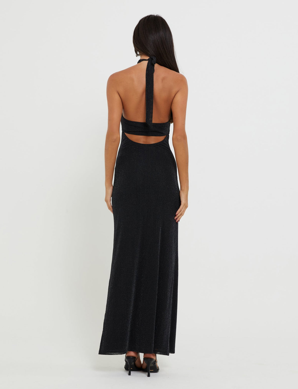 A woman with long dark hair stands facing away, showcasing the sleek, sleeveless TIANAH MAXI DRESS - BLACK from TIGERMIST. The halter neck gown reveals her back and features a cutout detail just below the shoulder blades. She pairs the floor-length dress with black heels and stands against a neutral background.