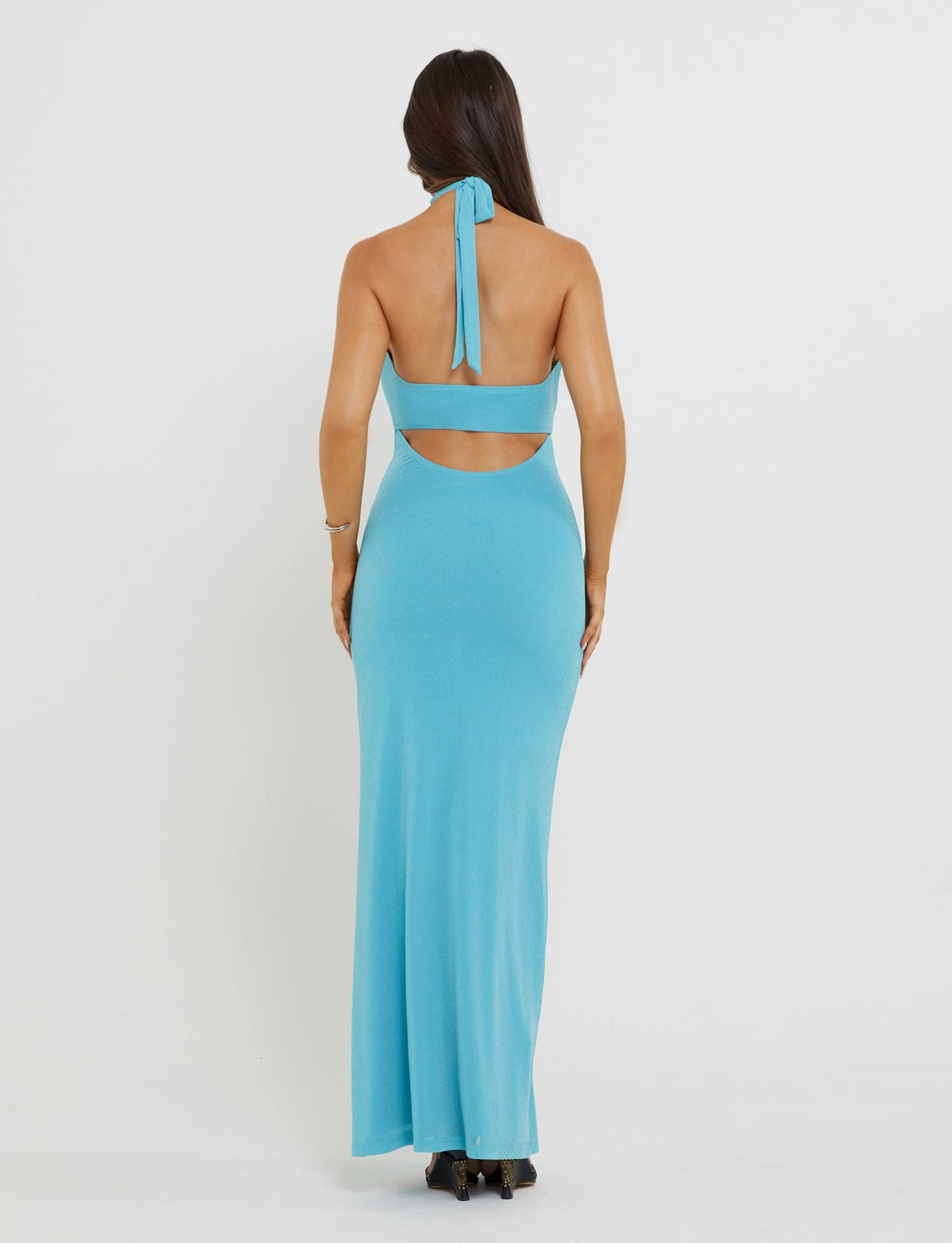 A woman is wearing the TIANAH MAXI DRESS in baby blue from TIGERMIST. She stands with her back to the camera, showing off the dress's tie closure and mid-back cutout. The floor-length dress glistens with blue sparkle as she pairs it with high-heeled shoes.