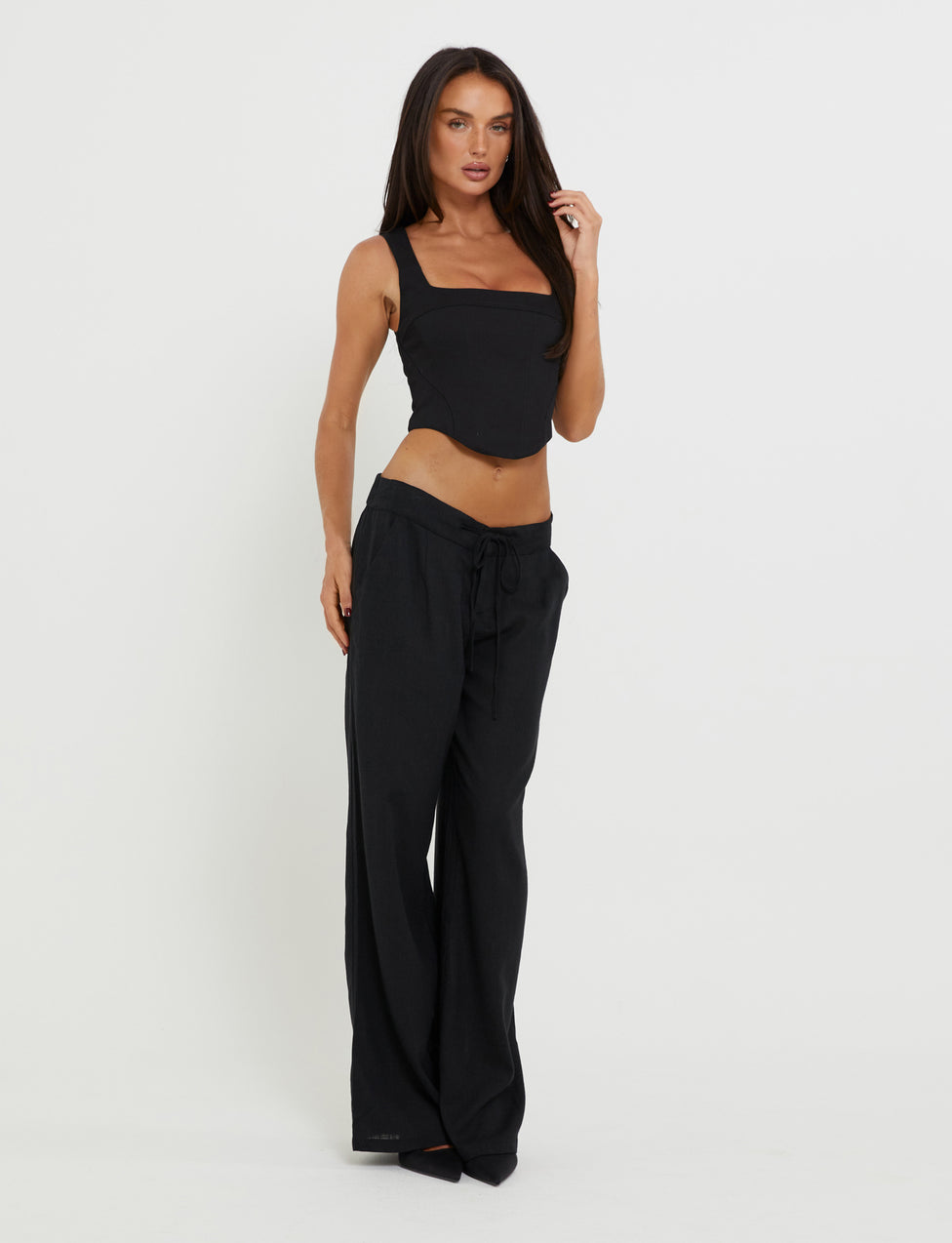 A woman with long dark hair, dressed in a TIGERMIST black sleeveless crop top and AIDEN PANT - BLACK, stands against a plain white background. One hand touches her hair while the other hangs by her side.