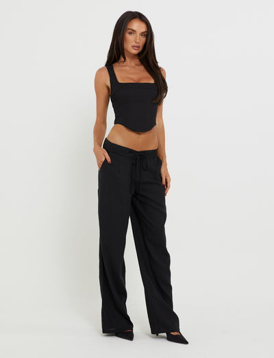 A woman stands against a plain white background, wearing a chic TIGERMIST SULLIVAN TOP - BLACK sleeveless square-neck top and high-waisted, wide-leg black pants with her hands in her pockets. She has long brown hair and is wearing black pointed-toe shoes.