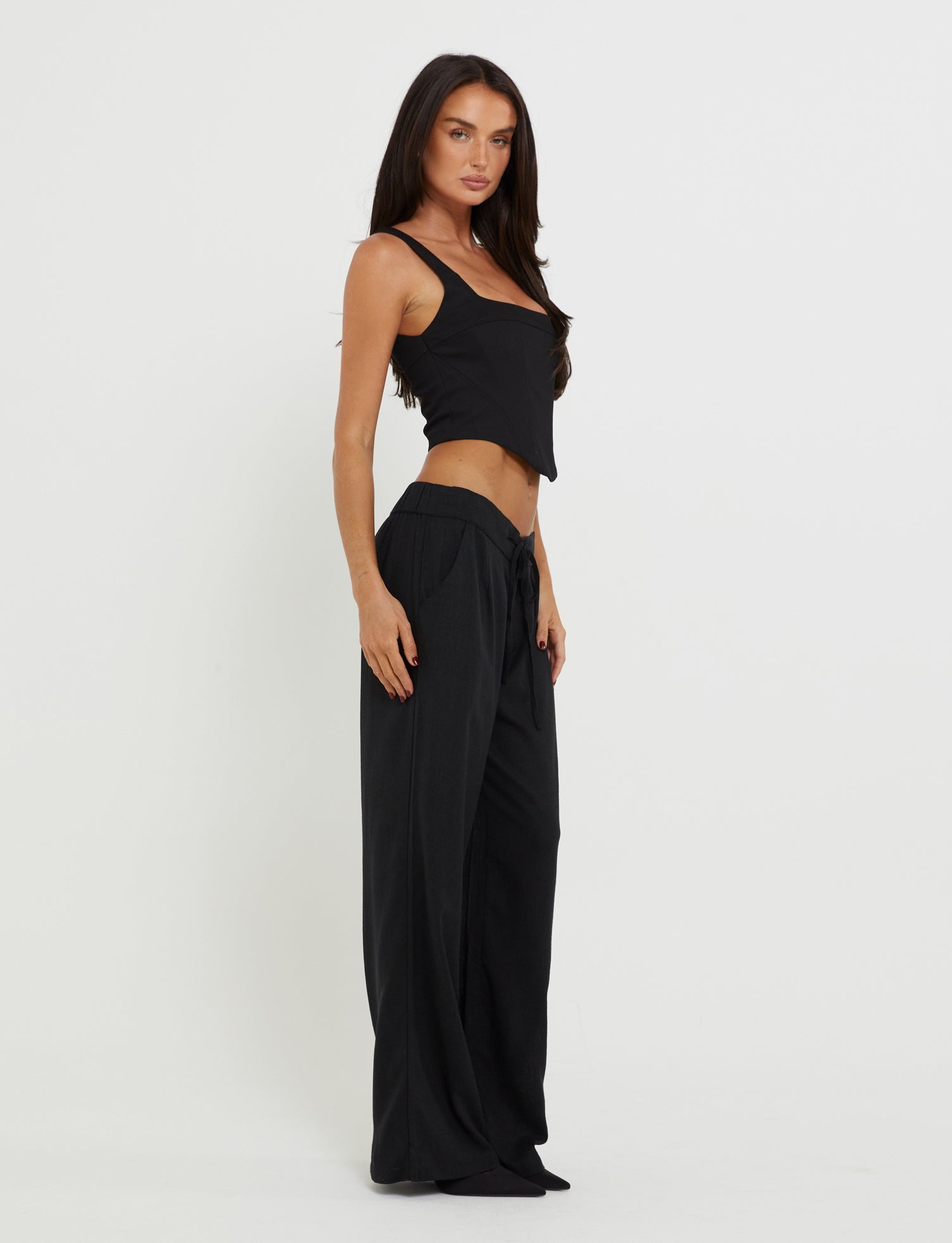 A woman poses against a white background, wearing the SULLIVAN TOP - BLACK from TIGERMIST and wide-legged pants. She boasts long dark hair and a serious expression, standing with one hand on her hip and slightly turned to her right.