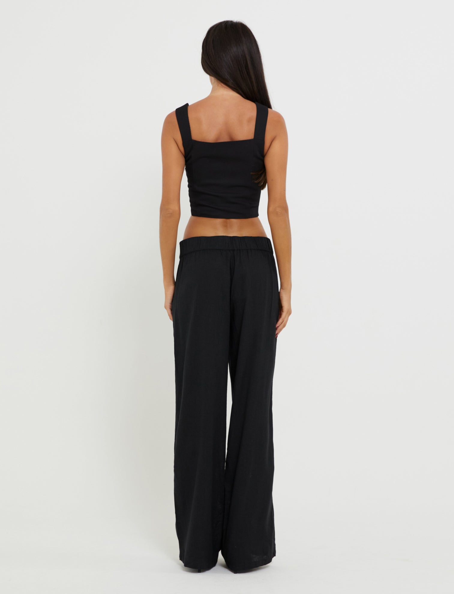 A woman with long dark hair, standing with her back to the camera, exudes confidence in her TIGERMIST SULLIVAN TOP - BLACK and matching wide-leg pants. The stark contrast of the outfit against the plain white wall enhances her striking silhouette.