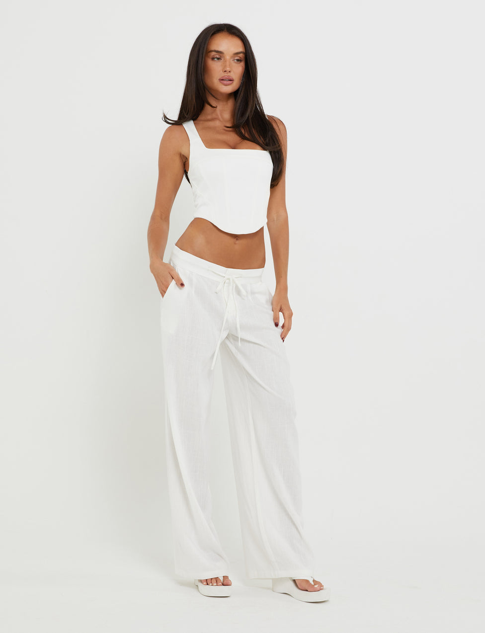A woman with long dark hair wearing a white sleeveless top and the AIDEN PANT - WHITE from TIGERMIST stands against a plain white background. She has her hands in her pants pockets and is looking slightly to the side. She is also wearing white flip-flops.