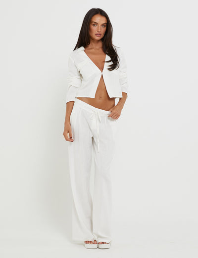 A woman stands against a plain white background, wearing the AIDEN TOP - WHITE by TIGERMIST and matching drawstring trousers. She has long brown hair and is looking directly at the camera with a neutral expression. She is barefoot.