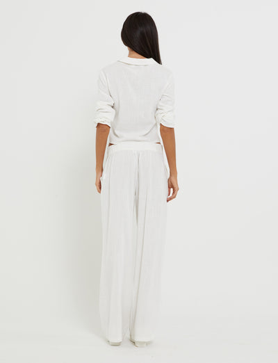 A person with long dark hair stands facing away, wearing the AIDEN TOP - WHITE from TIGERMIST paired with loose white pants. The outfit is relaxed and minimalist, featuring rolled-up sleeves and a slightly untucked hem. The background is completely white, creating a clean and simple look.