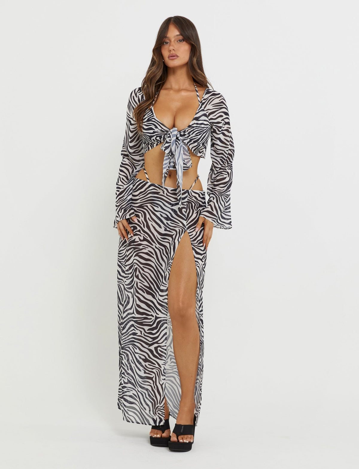 A woman stands against a plain white background wearing the TIGERMIST AYESHA SKIRT in black zebra print. The ensemble includes a long-sleeve, tie-front crop top paired with a high-slit maxi skirt. She has long hair, wears black platform heels, and exudes confidence.