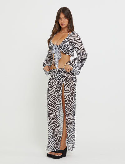 A woman is wearing the AYESHA TOP - BLACK : ZEBRA, a long-sleeve crop top by TIGERMIST, paired with a matching high-slit skirt. She is standing against a plain white background with her hands in the pockets of the skirt. Additionally, she is sporting black platform sandals.