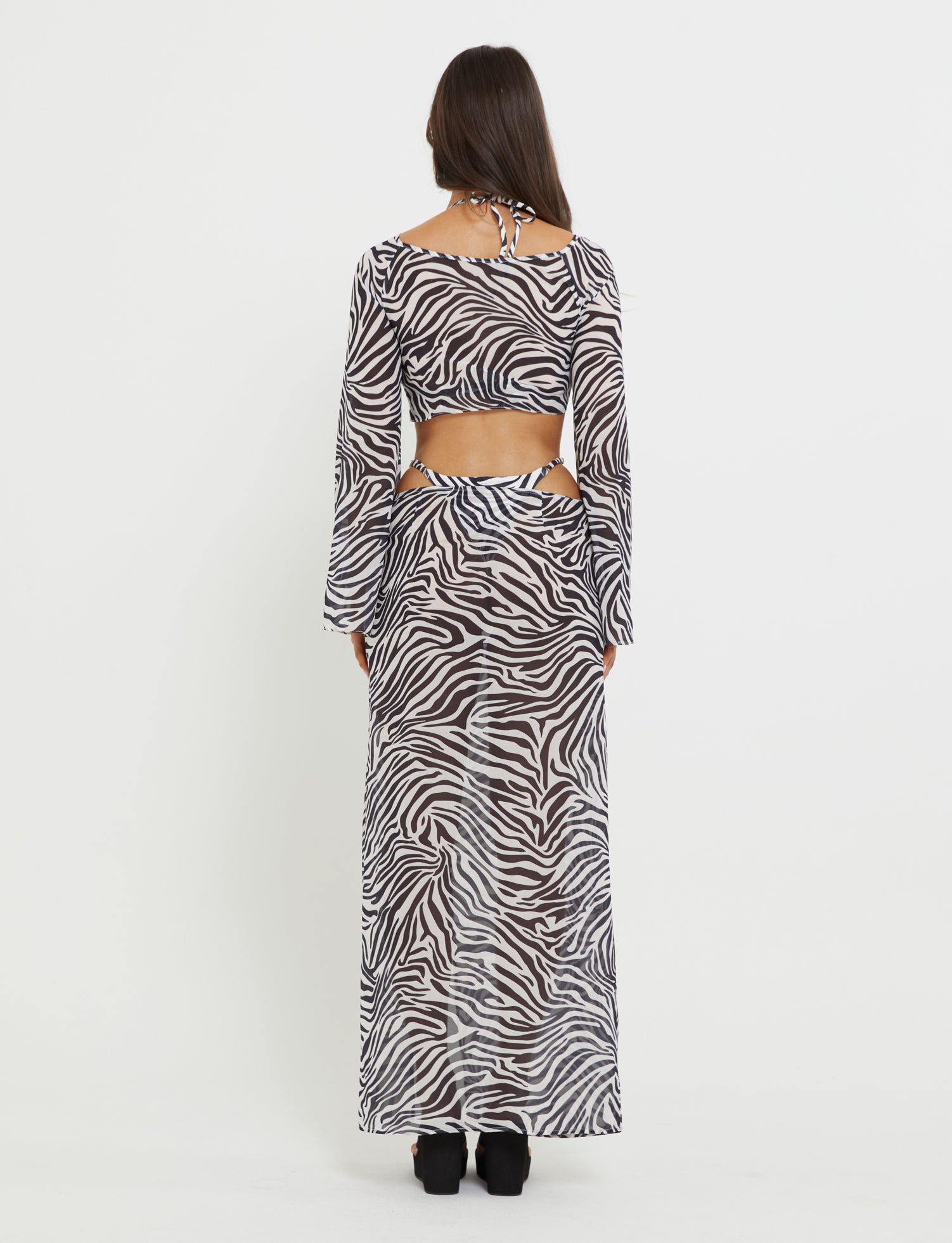 A person with long dark hair stands with their back to the camera, showcasing a Tiger Mist AYESHA Top in black zebra print. The stylish top features long sleeves and cutouts at the waist and back. They are wearing black shoes, set against a plain white background.