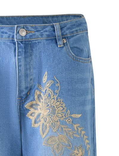 Close-up of a pair of VEGA JEANS in mid blue denim from TIGERMIST, featuring an intricate gold floral embroidery pattern on the upper left thigh area. The jeans have a button and zipper closure, along with standard belt loops and pockets.
