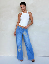 A person stands against a plain background wearing a fitted white tank top and the VEGA JEAN - BLUE from TIGERMIST. The mid-blue wide-leg jeans feature floral embroidery, and the person poses with one hand on their hip and the other resting on their thigh, looking directly at the camera. They complete the look with black open-toe heels.