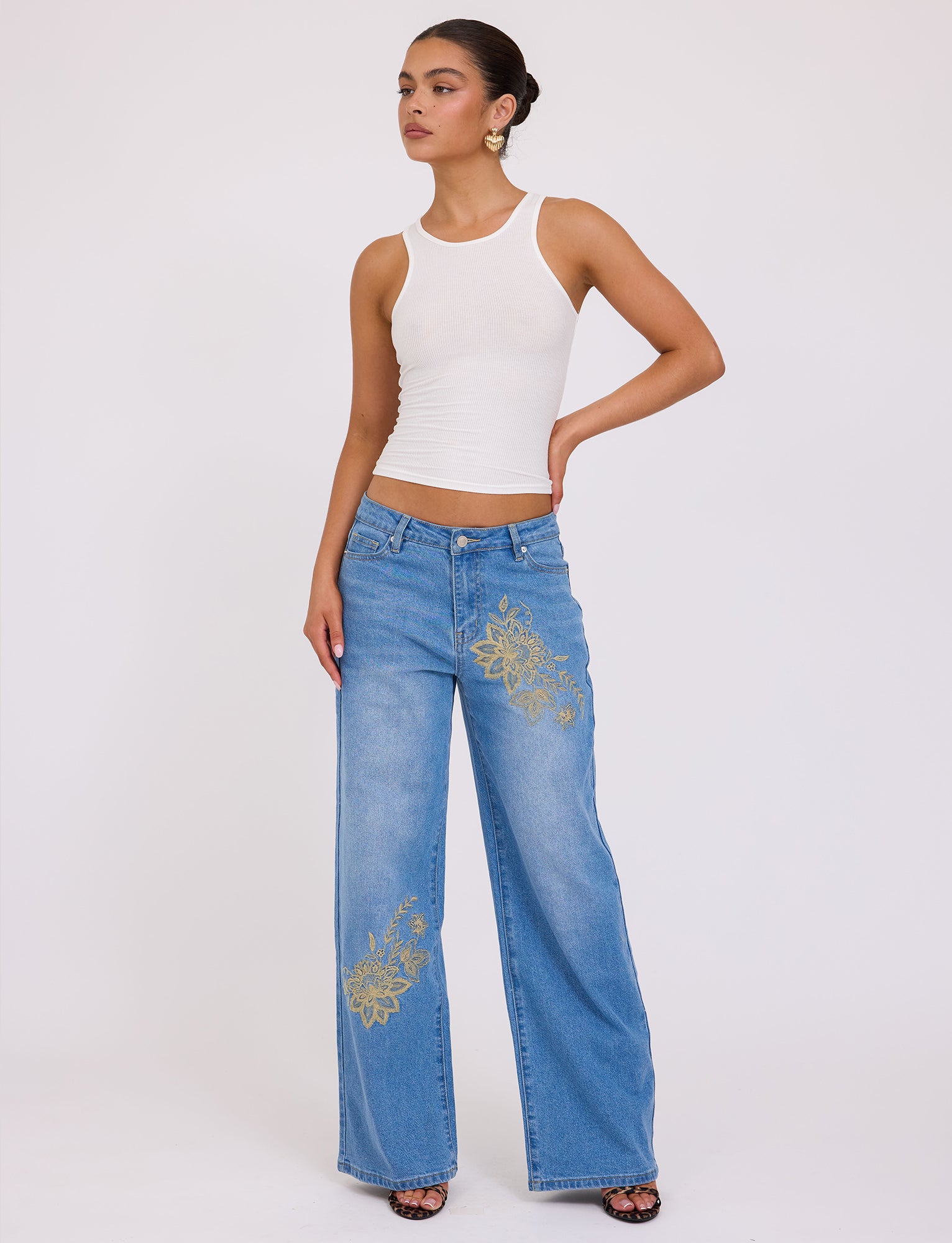 A person stands against a plain white background, wearing a white sleeveless top and the mid blue VEGA JEAN from TIGERMIST, featuring light blue wide-leg denim with floral embroidery on the left leg. They have their hair pulled back, accessorized with large hoop earrings, and are posing with one hand on their hip in this chic Tiger Mist ensemble.