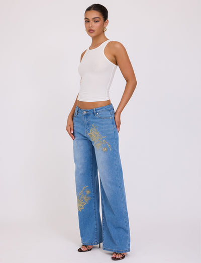 A woman stands against a plain white background wearing a sleeveless white top and the VEGA JEAN - BLUE in mid blue denim with floral embroidery on the left leg by TIGERMIST. She has gold hoop earrings and black sandals. Her hair is pulled back.