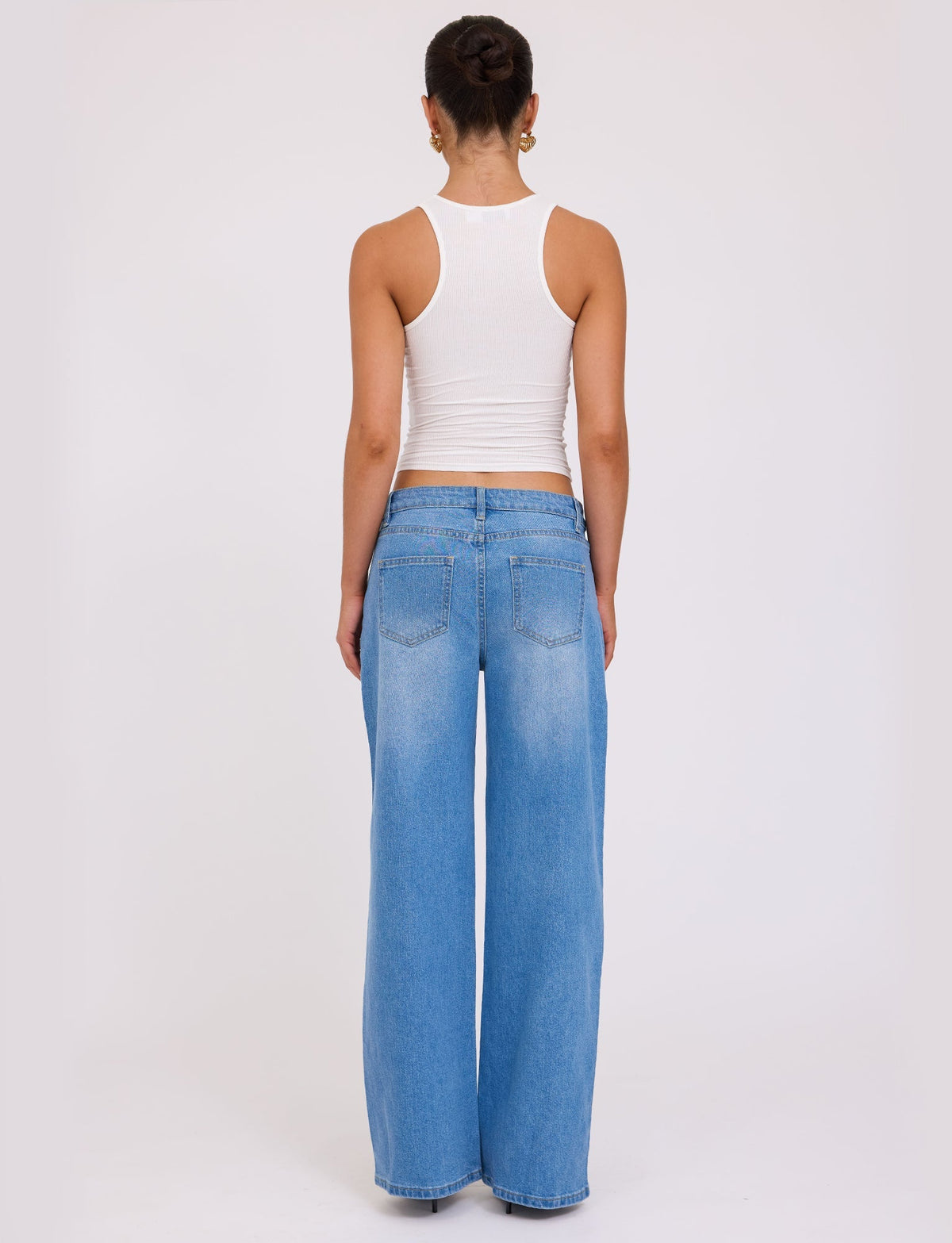 A person with a dark hair bun is standing with their back to the camera, wearing a white sleeveless top and the VEGA JEAN - BLUE : DENIM : MID BLUE from TIGERMIST. The background is plain and white, providing a clean and simple setting.